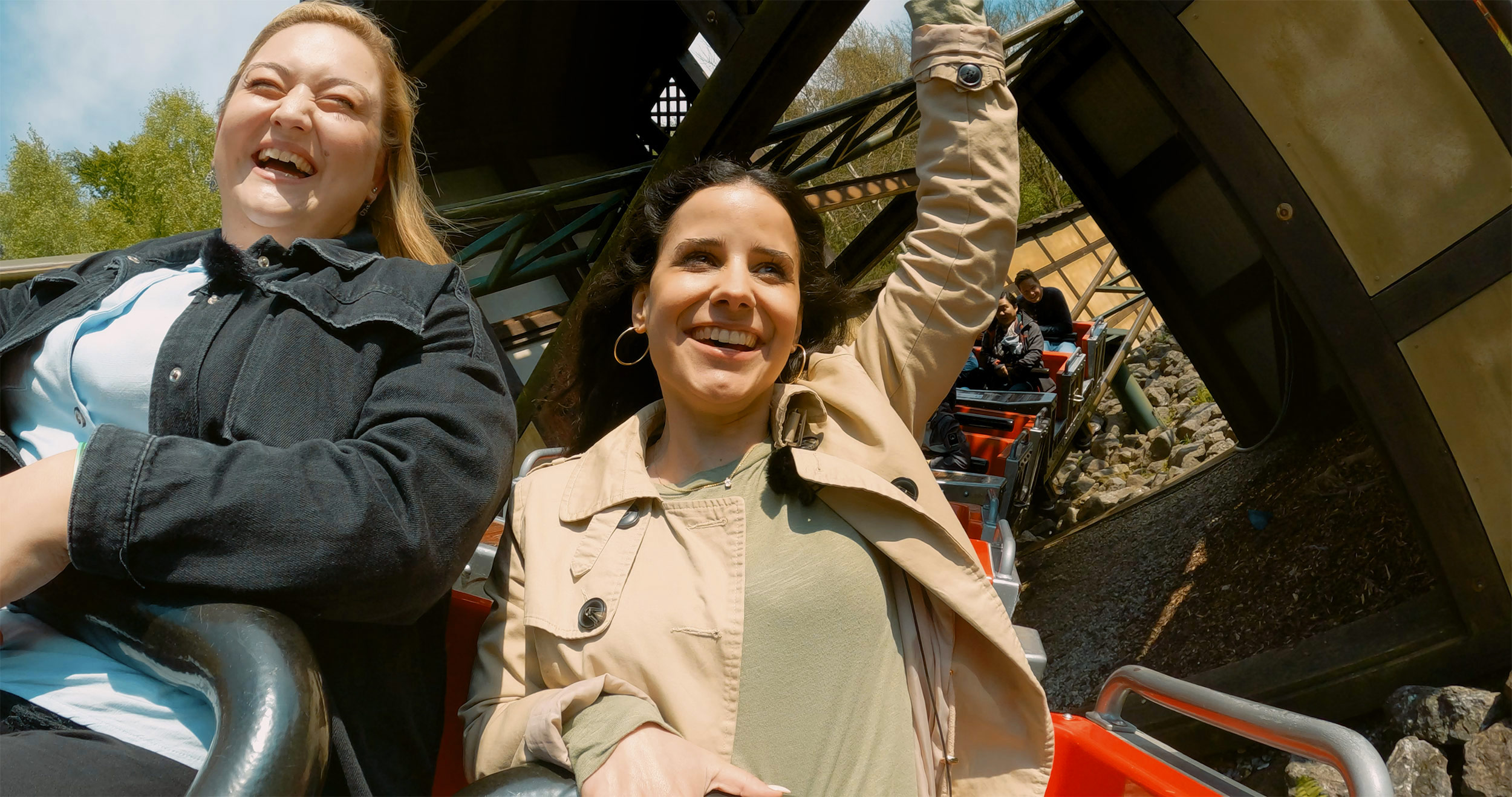 Zwei Frauen, die in der Achterbahn "Drachenjagd" des LEGOLAND Deutschlands sitzen und lachen, wobei die Frau rechts ihre rechte Hand festhält und ihre linke Hand in die Luft hebt, während im Hintergrund die Wagen des Fahrgeschäfts und die Schienen der Achterbahn zu sehen sind.